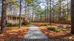 A small house at 0 Dutchess Lane with a front porch sits in a wooded yard in Henderson, surrounded by tall pine trees and a wooden fence. A gravel path leads to the house, with landscaped plants and mulched ground throughout.