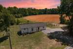 A small white house with a fenced yard at 4004 Old NC 75 in Stem sits next to a large, freshly plowed field bordered by trees under a vibrant pink and orange sunset sky.