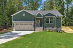 A green single-story house with white trim at 114 Comanche Drive, Louisburg, features a two-car garage, small front porch, and sits on a well-manicured lawn surrounded by tall trees and a concrete driveway.
