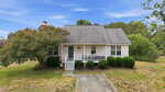 A small white house at 15 Magnolia Ridge Drive, Louisburg, features a front porch with steps and railing, partially shaded by a large tree. Sparse grass and bushes fill the yard, while an American flag hangs by the doorway under a blue sky.