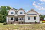 Two-story modern farmhouse at 1206 Pond Side Drive in Creedmoor features white siding, brown trim, a covered front porch, brick foundation, and manicured landscaping on a grassy lawn with trees and blue sky in the background.