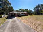 Charming single-story red brick house at 300 7th Street, Butner, with a covered carport, welcoming front porch, large grassy yard, and paved driveway. Mature trees surround the property on a sunny day.