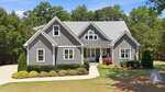Nestled in Wake Forest, this modern gray house with white trim, gabled roofs, and a covered front porch at 3981 Hope Valley Drive sits amid green trees and manicured shrubs. A curved driveway welcomes you to the entrance under a blue sky.