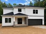 A modern two-story white house with black trim at 107 Acoma Drive, Louisburg, features a covered front porch with stone accents, a dark double garage door, and large windows, set on a dirt lot surrounded by trees under a blue sky.