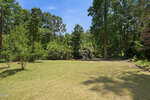A grassy backyard at 126 Wanna Drive in Louisburg, bordered by tall green trees and shrubs under a clear blue sky, with sunlight casting shadows across the lawn.