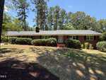 A single-story brick house with white trim at 174 Pine Cone Drive, Oxford, nestles among bushes and tall pine trees under a clear blue sky. The front yard is partly shaded, with grassy patches and a brick walkway leading to the door.