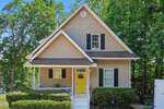 A beige two-story house at 383 Sagamore Drive, Louisburg, with white trim, black shutters, and a bright yellow front door. Green bushes and tall trees surround the small porch with white railings and steps leading to the entrance.
