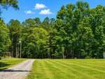 A gravel driveway at 0 Huntsboro Road curves through a neatly mowed green lawn in Oxford, leading toward a dense forest of tall, leafy trees under a bright blue sky with a few small clouds.