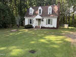 A white, two-story house with dormer windows and a brown roof sits on a large, grassy yard at 964 Hicks Road in Youngsville. A fire pit and circular patch are visible on the lawn, with steps leading up to the front door.