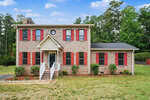 Charming two-story brick house at 603 20th Street in Butner, featuring red shutters and a covered front entrance with white railings. The grassy yard is dotted with small shrubs, and mature trees stand under a partly cloudy sky.