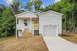 A modern white house in Louisburg with a yellow front door, single-car garage, wood steps to a cozy porch, and a concrete driveway at 405 Ridley Street, surrounded by trees and a patchy lawn.