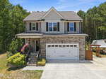 Charming two-story house at 20 Mitchell Ridge Drive in Youngsville, featuring stone and siding exterior, black shutters, a covered front porch with American flags. The driveway leads to a double garage; trees and a boat are visible in the background.