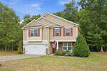 A two-story beige house with red shutters at 1842 Fillmore Drive, Creedmoor, featuring a white garage door, an American flag by the entrance, and a driveway, all surrounded by green trees and a grassy yard under a blue sky.