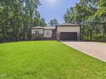 A modern single-story house at 132 Mohawk Drive, Louisburg, with a stone and siding exterior, black garage door, and a wide driveway, surrounded by a large, neatly mowed lawn and tall trees under a clear blue sky.