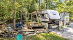 A white Kodiak travel trailer is parked at a wooded Louisburg campsite on 171 Winnebago Loop, with potted plants, outdoor seating, a hammock, and a small shed nearby. Sunlight filters through the trees, creating a peaceful atmosphere.