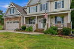 Charming two-story suburban home at 3535 Lavender Lane, Wake Forest, with stone and siding exterior, front porch with white railings, hanging plants, an American flag, and attached two-car garage. Well-kept lawn and shrubbery enhance the curb appeal.