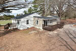 Single-story house at 2105 Newton Dairy Road, Henderson, featuring beige and white siding, two wooden decks, and surrounded by leafless trees. The yard is mostly bare dirt, with a gravel driveway visible to the right.