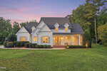 A charming white house with dormer windows at 335 Forest Bridge Road in Youngsville features a covered front porch and glowing interior lights, set on a large, well-manicured lawn with trees in the background at sunset.