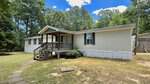 Charming single-story beige manufactured home at 10608 Hicksboro Road, Oxford, featuring a small covered front porch with wooden steps and white trim, nestled among trees and grass beneath a blue sky with scattered clouds.