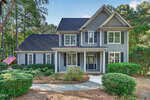Charming two-story gray house with white trim and black shutters at 210 Longwood Drive, Youngsville. Surrounded by green shrubs and tall trees, an American flag adorns the front as a concrete pathway leads to the welcoming porch.