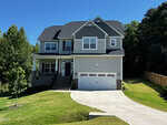 Two-story modern suburban house at 5 Cinnamon Teal Way, Youngsville, featuring gray siding, a covered porch, black roof, and a white two-car garage. Set on a sloped lawn with curved driveway, trees, and a wooden fence.