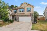 Charming two-story suburban house at 721 Tannerwell Avenue, Wake Forest, with beige siding, stone accent facade, double garage, and basketball hoop in the driveway; trimmed bushes and a small tree highlight the front yard amid neighboring homes.