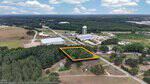 Aerial view of a wooded lot at 0 Bear Pond Road outlined in yellow within a semi-rural Henderson area. Nearby are houses, trees, roads, large industrial buildings, and a tall white water tower in the background.