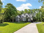 A large suburban house at 1006 Canterbury Court, Youngsville, features a gray stone and siding exterior, surrounded by green lawn, trees, and shrubs, with a long concrete driveway leading to the front entrance under a blue sky with scattered clouds.