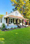 A charming white house with dark shutters at 2763 Rock Mill Road, Henderson, features a large front porch with wooden columns, rocking chairs, and a quilt-pattern flag. It's surrounded by lush grass, vibrant plants, and tall trees beneath a clear blue sky.