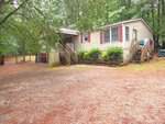 A beige single-story house at 3147 Buckhorn Lane, Wake Forest, features a front porch, two sets of steps, and shrubs along the front, surrounded by tall trees and a dirt driveway in a wooded area.