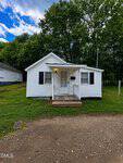 Charming small white house with black shutters and a front porch at 412 Brick Street in Henderson, set on a grassy lot with tall green trees behind and a gravel driveway running out front.