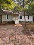 A small, light-colored house with white trim, shutters, and a front porch sits among tall trees at 113 Cochise Drive in Louisburg. A gravel path leads to the steps, with simple landscaping featuring shrubs and mulch beds in the yard.