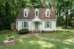 A charming white two-story house with dormer windows and green shutters sits surrounded by grass and trees, featuring a small porch, garden path, birdbath, and shed in the backyard.