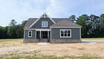 A modern gray house with white trim at 20 White Deer Lane, Youngsville, features a stone foundation and a peaked roof on a dirt lot with a partial concrete driveway, surrounded by trees under a blue sky.