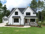 A modern farmhouse-style home at 220 Whistlers Cove in Louisburg, featuring white board-and-batten siding, black window frames, a covered front porch with stone accents, a detached garage, green lawn, and tall trees.