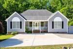 Single-story gray house with white trim, black shutters, a covered front porch, and large arched windows at 1608 Rogers Pointe Lane in Creedmoor, set against a backdrop of trees with a wide driveway in front.