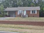 A single-story brick house with white trim at 109 Moss Road in Creedmoor, featuring a small front porch and attached carport, set on a grassy lawn with a low brick wall by the roadside. Pine trees line the background.