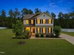 Charming two-story house at 185 Green Haven Boulevard, Youngsville, features lit windows at dusk, dark shutters, a manicured lawn, shrubs, young trees, and a long driveway set against tall trees and a white fence.