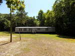 A long, single-story gray mobile home sits on a grassy, partially shaded lot at 0 E River Road in Louisburg, surrounded by tall green trees under a clear blue sky. Wooden steps lead to the front door.