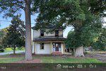 A two-story white house at 12670 Robbins Road in Youngsville features a covered porch and red front door, surrounded by large trees. Sunlight filters through the branches onto this charming, older home with 2585 sq ft, 3 beds, and 2 baths.