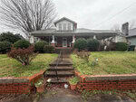 A two-story house at 414 Raleigh Road, Oxford, with a wide front porch, brick steps, and railing leading up to the entrance. Bushes and grass line the walkway. The sky is overcast and trees surround the house.