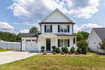 A two-story white house with black shutters at 101 Averly Court, Stem features a covered front porch, attached garage, and an American flag. The manicured lawn, neat bushes, and paved driveway complete the charming curb appeal under a partly cloudy sky.
