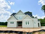 A newly constructed light gray house with white trim and a dark roof stands on bare, sandy ground at 215 Broadleaf Lane, Youngsville. Some windows still have stickers. Blue sky and trees are seen in the background.