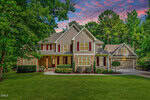 A two-story suburban house at 1006 Chatham Court in Youngsville features a stone and siding exterior, red shutters, covered porch, manicured lawn, double garage, and is surrounded by lush trees under a colorful sunset sky.