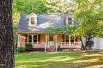 A beige, two-story house with a front porch, red shutters, and a sloped roof sits amid green trees and a grassy yard at 709 Appaloosa Way, Wake Forest, on a sunny day.