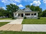 A single-story house at 202 Forest Avenue, Oxford, features a wide front porch with white railings and a gray-green exterior. Set on a large grassy yard under blue skies, the home is surrounded by trees with a driveway and sidewalk leading up.