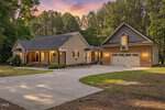 A tan single-story house at 160 Baron Circle, Youngsville, features a front porch with an American flag, an attached two-car garage, and a curved driveway set among tall trees at sunset.