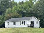 A small, single-story gray house with white trim sits on a grassy lawn at 695 Layton Store Road in Castalia, surrounded by dense green trees. The house has several windows and a small front porch with steps leading up to the door.