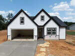 A newly constructed white house with black trim at 6218 Carole Drive, Oxford, features a double garage and an unfinished yard with dirt and scattered wooden boards, all beneath a blue sky with scattered clouds.