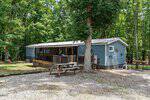 A blue single-story house at 111 Nature Lane in Henderson features a screened porch, surrounded by tall trees and a gravel driveway. A picnic table, grill, and outdoor seating sit in the shaded yard as sunlight filters through the forested background.